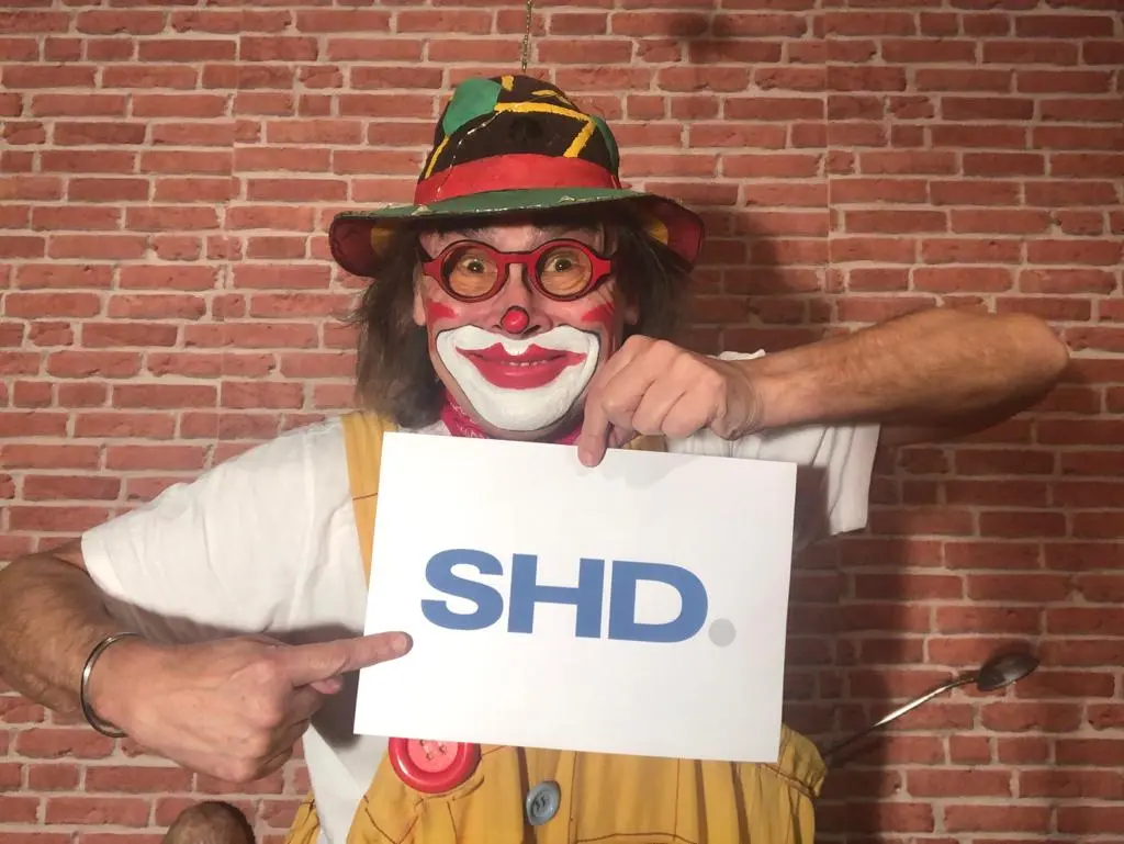 Ein bunt bemalter Clown hält das SHD Logo nach oben und zeigt darauf.