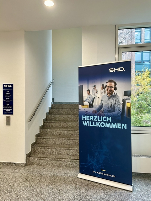 Ein Begrüßungsbanner von SHD steht in einem hellen Treppenhaus mit Informationen zur IT-Dienstleistung.
