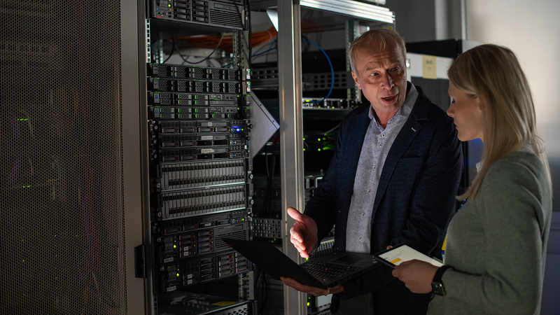 Zwei IT-Fachkräfte diskutieren vor einem Server-Rack in einem Rechenzentrum, verwenden Laptop und Tablet, moderne Server-Hardware und Netzwerkinfrastruktur im Hintergrund.