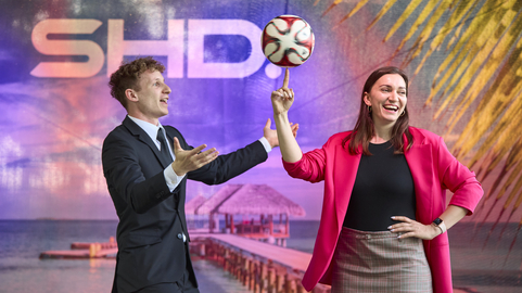 Ein Mann im Anzug und eine Frau in bunter Kleidung spielern mit einem Fußball vor einem SHD-Hintergrund.
