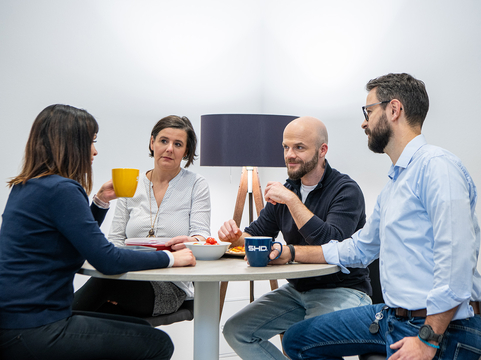 In der Mittagspause unterhalten sich vier Mitarbeitende von SHD beim Essen an einem runden Tisch.
