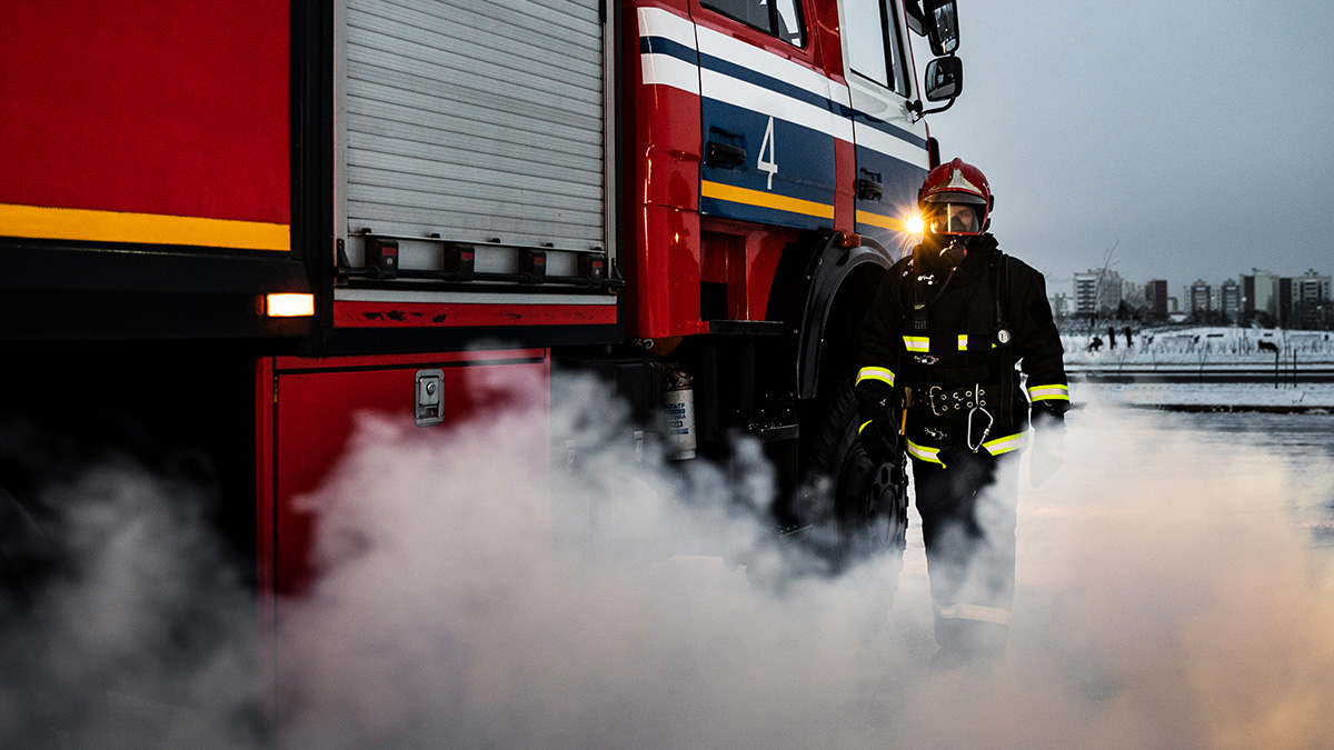Ein Feuerwehrmann in voller Ausstattung läuft um ein Löschfahrzeug. Um ihn herum ist Rauch und roter Schein von einem Feuer in der Nähe.