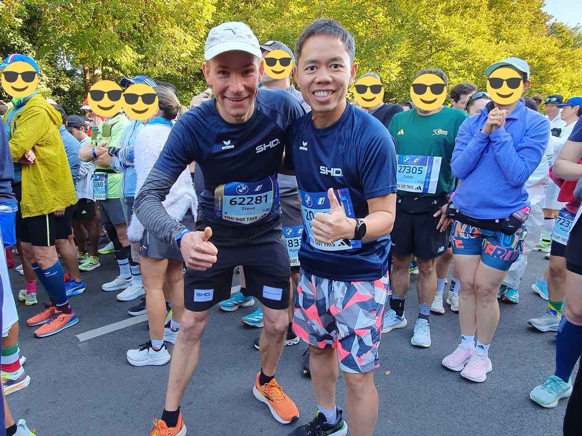 Zwei Männer in SHD-Shirts posieren begeistert vor einem Lauf, umgeben von Teilnehmern und Bäumen.