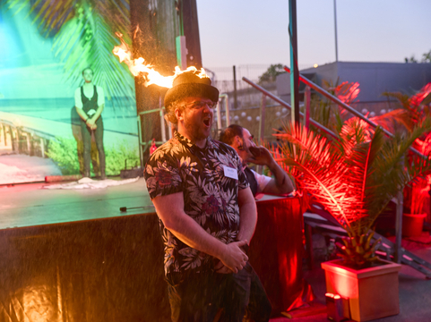 Ein Mann mit Feuerhut singt leidenschaftlich auf einer bunten Bühne bei einer SHD-Veranstaltung.