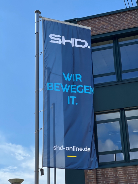 Flagge mit dem Schriftzug "SHD. WIR BEWEGEN IT." vor dem modernen Bürogebäude der Dresdner Geschäftsstelle.