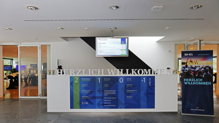 Empfangstresen mit Willkommenstext, Informationsdisplay und SHD-Bannern im modernen Konferenzraum.