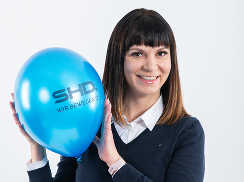 Eine Person hält einen blauen Ballon mit dem Logo von SHD und lächelt freundlich.
