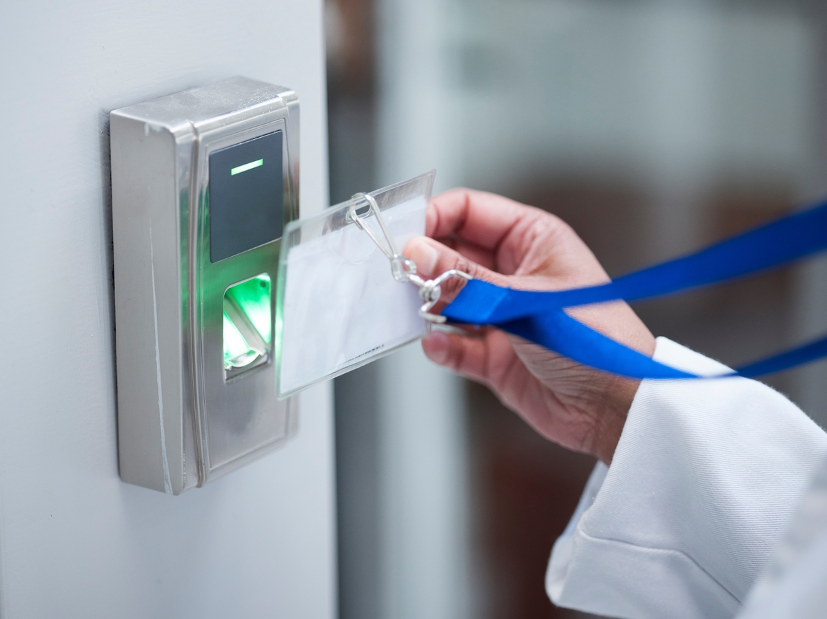 Eine Hand im weißen Kittel als Symbol für KRITIS-Angestellte hält ihren Ausweis mit Lanyard in einen Scanner an der Wand.