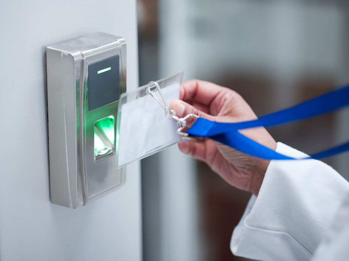 Eine Hand im weißen Kittel als Symbol für KRITIS-Angestellte hält ihren Ausweis mit Lanyard in einen Scanner an der Wand.
