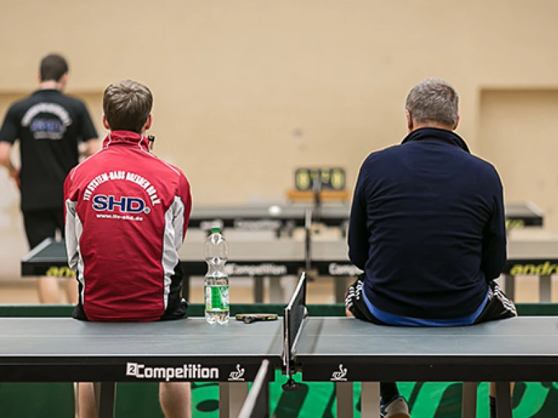 Zwei Männer sitzen entspannt auf einer Tischtennisplatte, einer trägt eine rote SHD Trainingsjacke