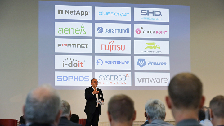 Ein Referent spricht vor einem Publikum, während Logos von IT-Partnern wie SHD angezeigt werden.