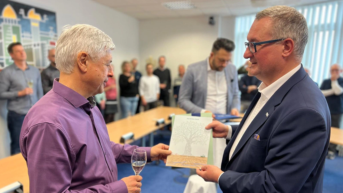 SHD Geschäftsführer übergeben Dr. Frank Karow eine Karte, im Hintergrund feiern mehrere Personen mit Getränken.