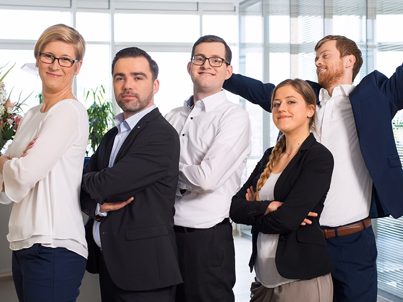 Lächelnde Mitarbeitende von SHD in einer Reihe schauen in die Kamera, einer streckt sich fröhlich