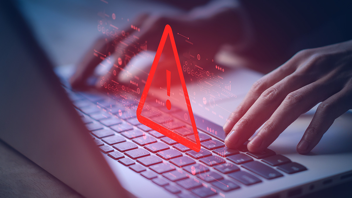 Zwei Hände bedienen einen Laptop, über der Tastatur leuchtet ein rotes Warndreieck auf.