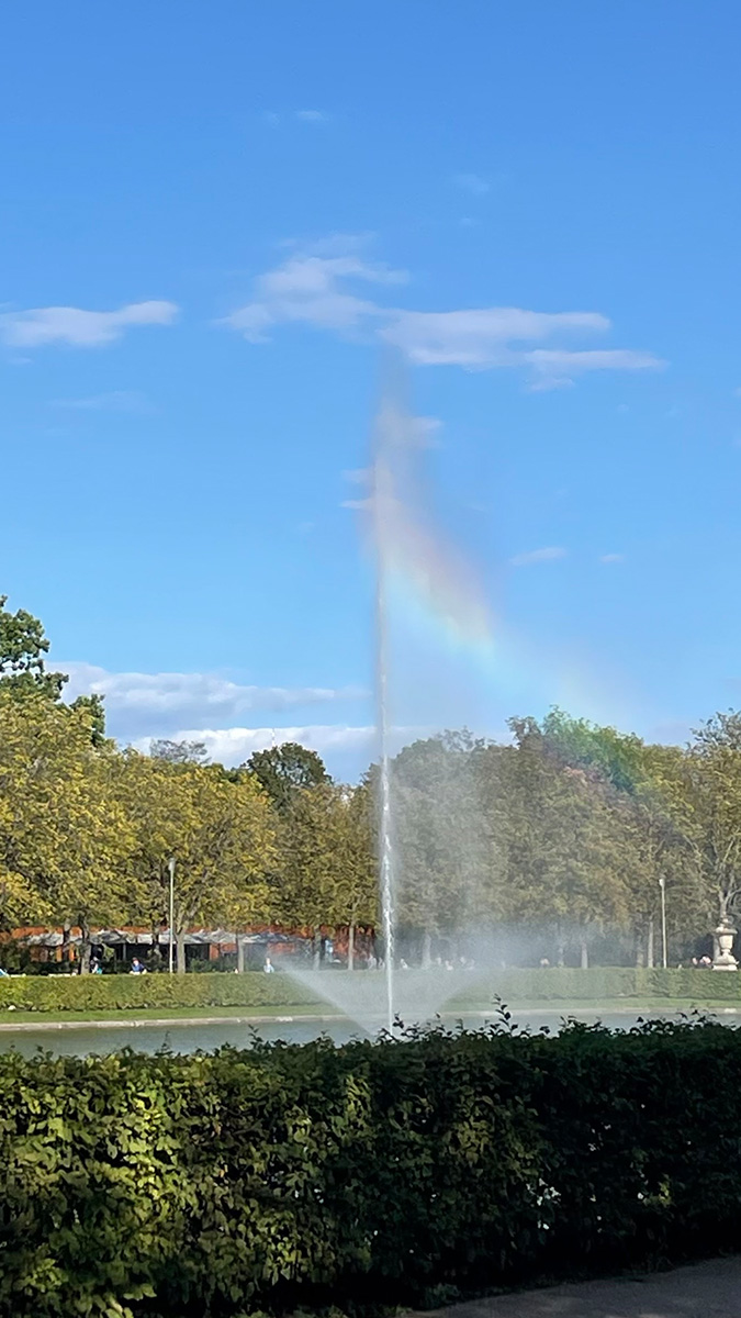 Springbrunnen mit Regenbogen am Palais Großer Garten in Dresden vor einem blauen Himmel und grünen Bäumen.
