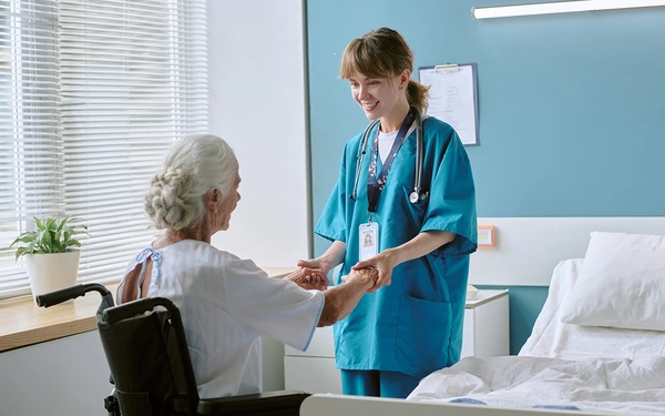 Eine junge lächelnde Krankenschwester kümmert sich um eine alte Damen im Rollstuhl in einem modernen Krankenzimmer.