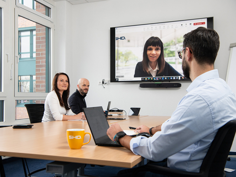 SHD Mitarbeitende dikutieren in einem hybriden Meeting über ein aktuellen Projekt. Ein Teil sitzt am Konferenztisch und eine Kollegin ist remote am Monitor dabei.
