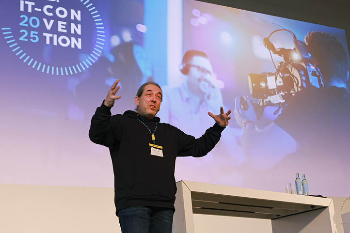 Ein Redner erklärt innovative IT-Lösungen auf einer Konferenz, während eine Kamera filmt. SHD fördert digitale Transformation.