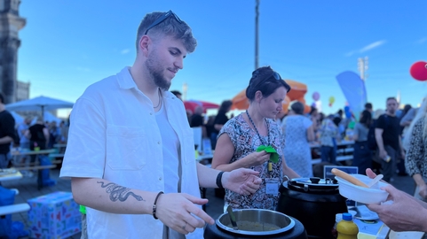 Ein junger Mann serviert Essen an einem bunten Fest mit vielen Besuchern und Ständen.