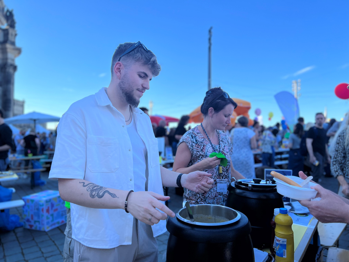 Ein junger Mann serviert Essen an einem bunten Fest mit vielen Besuchern und Ständen.