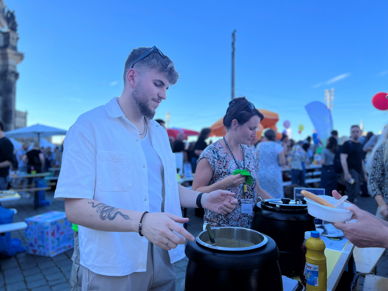 Ein junger Mann serviert Essen an einem bunten Fest mit vielen Besuchern und Ständen.