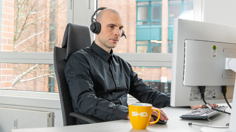 Ein IT-Mitarbeiter von SHD arbeitet am Computer mit Headset und Kaffeetasse im Büro.