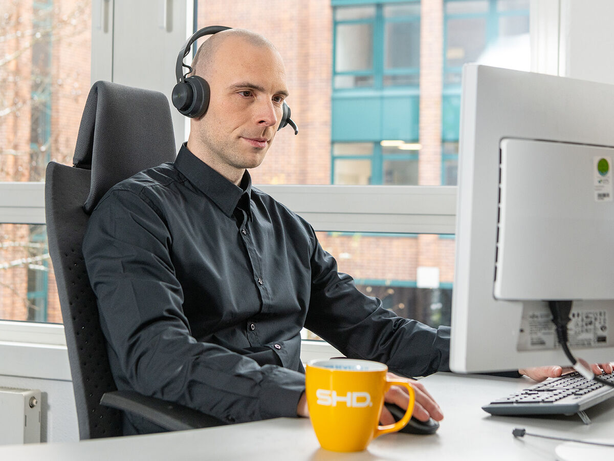 Ein IT-Mitarbeiter von SHD arbeitet am Computer mit Headset und Kaffeetasse im Büro.