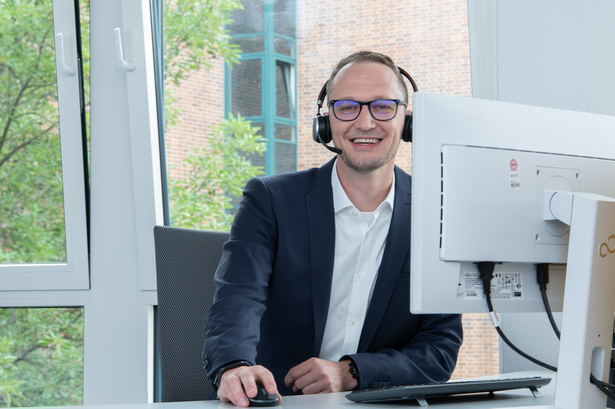 Ein Mann mit Headset lächelt und nutzt einen Computer in einem modernen Büro.