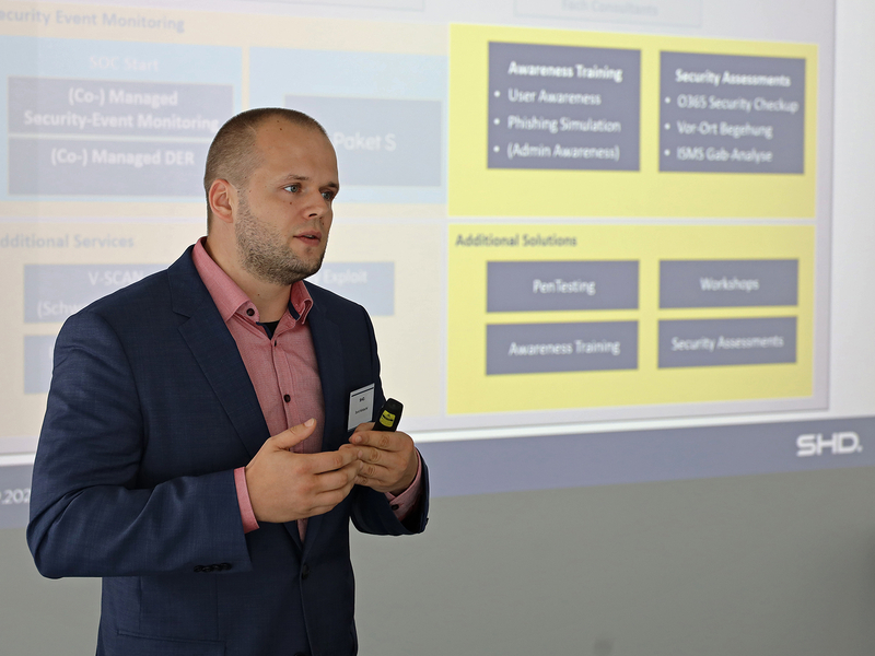 Ein Mann präsentiert IT-Sicherheitslösungen von SHD, während eine Präsentation im Hintergrund läuft.
