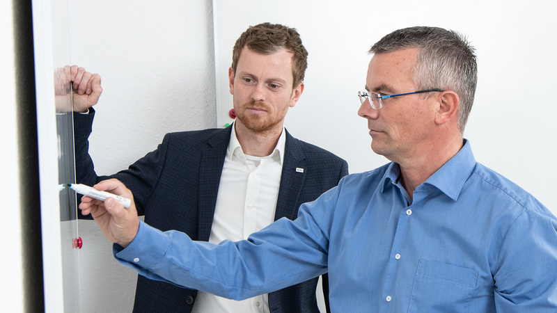 IT-Experte von SHD bespricht ein Projekt mit seinem Kollegen an einem Whiteboard und schreibt mit einem Marker.