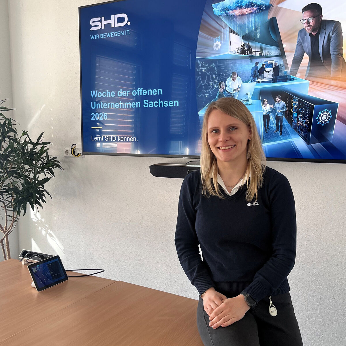 Die Ausbildungskoordinatorin von SHD lehnt am Tisch in einem Meetingraum und lächelt, im Hintergrund ein Monitor mit der Überschrift: Woche der offenen Unternehmen Sachsen 2026.