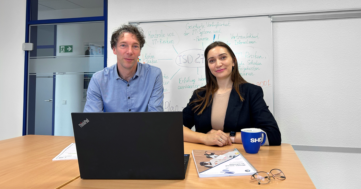 Zwei Personen sitzen an einem Tisch, mit Laptop und Dokumenten, in einem Büroumfeld.