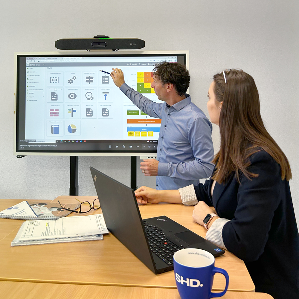 Ein Mann zeigt auf einen Bildschirm, während eine Frau aufmerksam zuhört, in einem Besprechungsraum.