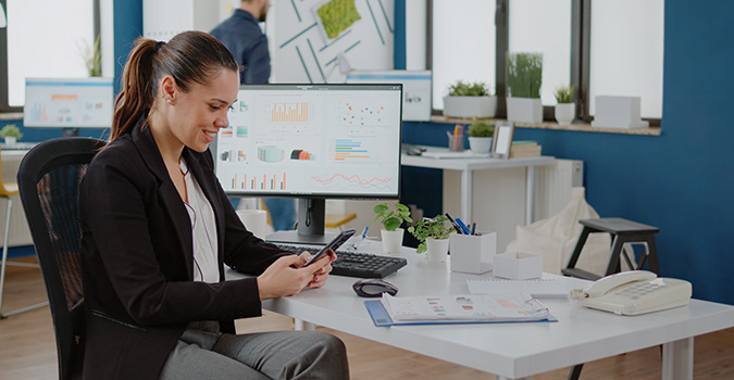 Eine Frau sitzend mit dem Smartphone in einer modernen Büroumgebung, Computer mit Diagrammen im Hintergrund.