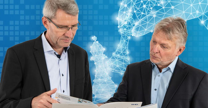 Zwei Männer betrachten gemeinsam ein Heft, während im Hintergrund Schachfiguren bestehen.