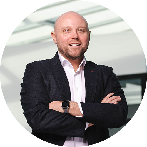 Mann im Anzug steht selbstbewusst mit verschränkten Armen in modernem Büro-Umfeld, repräsentiert SHD.