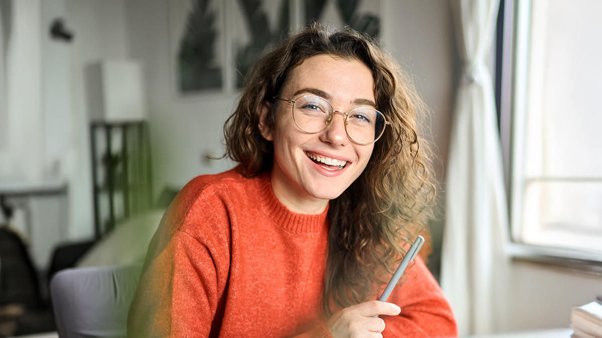 Junge Studentin mit Brille sitzt am Schreibtisch und lächelt fröhlich in die Kamera.