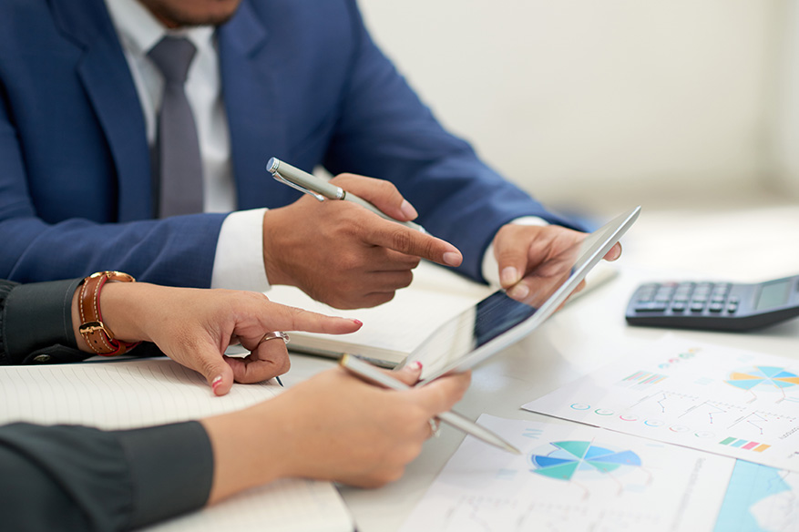 Die Hände von zwei Personen in einer Consulting-Situation mit Tablet, auf dem Tisch liegen Dokumente, Notizbücher und ein Taschenrechner